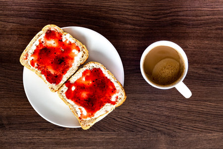 Top view on a plate with breakfast, two slices of bread with fruit marmalade, hot coffee on a dark wooden deskの写真素材