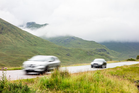 Two cars driving on a road in the landscape of Scottish Highlands in misty weather with motion blur...