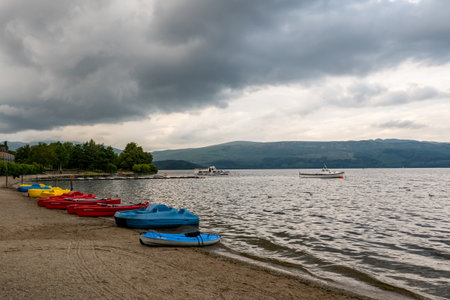 LUSS, SCOTLAND - AUGUST 1, 2019: Kayaks and other boats on a shore of the Loch Lomond in Scotland with a cruise ship in the backgroundのeditorial素材
