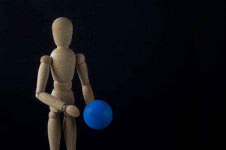 A wooden dummy holds a blue ball. Black background, close-up, copy space, horizontal orientation.の写真素材
