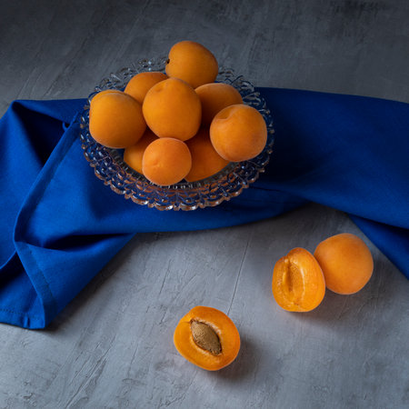 Apricots are lying in a transparent glass bowl, standing on a gray concrete background. Next to it is a blue cloth. Square orientation.の写真素材