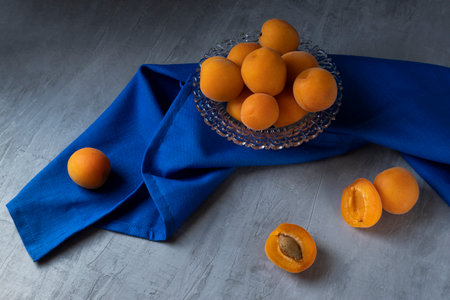 Apricots are lying in a transparent glass bowl, standing on a gray concrete background. Next to it is a blue cloth. Horizontal orientation.の写真素材