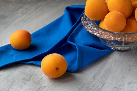 Apricots lie in a transparent glass bowl, standing on a gray concrete background. Next to it is a blue cloth. Close-up.の写真素材