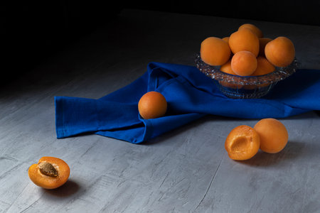 Apricots are lying in a transparent glass bowl, standing on a gray concrete background. Next to it is a blue cloth.の写真素材