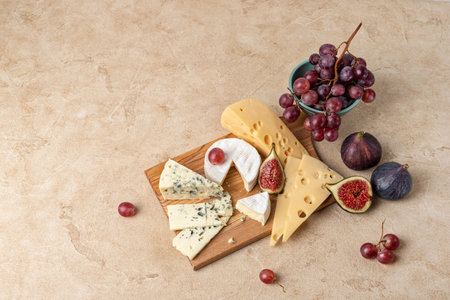 Maasdam cheese, camembert and blue cheese are served as an appetizer with fruit. Several types of cheese lie on a wooden board surrounded by figs and grapes. Top view, beige background, copy space.の写真素材