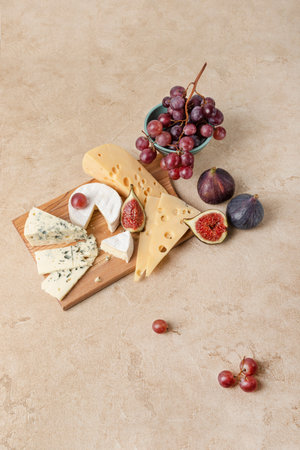 Several types of cheese lie on a wooden board surrounded by figs and grapes. Maasdam cheese, camembert and blue cheese are served as an appetizer with fruit. Top view, beige background, copy space.の写真素材