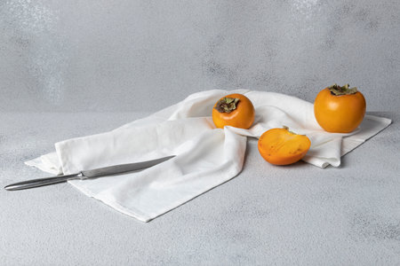 Still life with persimmon. The fruits are lying on a white towel, next to them is a knife. Gray textured background, horizontal orientation, copy space.の写真素材