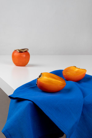 A cut ripe persimmon is lying on a blue towel. White table, close-up, vertical orientation and copy space.の写真素材