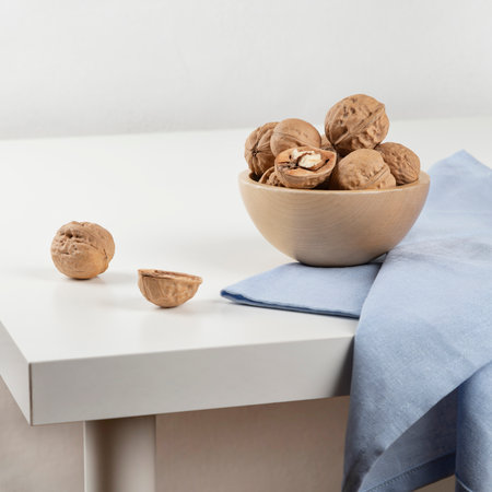 There is a wooden bowl with walnuts on a white table, and a blue towel is lying next to it....