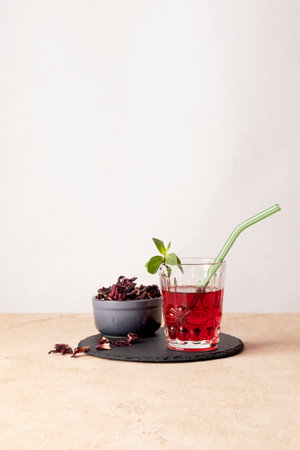 Karkade tea (hibiscus) in a glass with a glass tube and mint leaves. There is a ceramic bowl with dry tea nearby.の写真素材