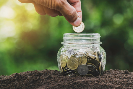 Hand of person holding a coin putting in glass jar in nature background to save money for the future...