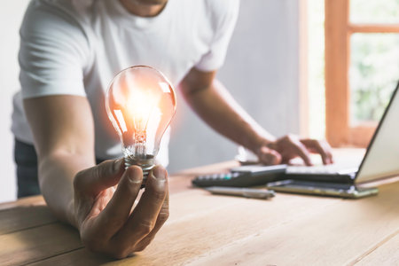 Business man holding light bulb on the desk in office and using  computer in  financial,accounting,energy,idea concept.の写真素材