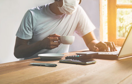 Man working at home in covid 19 virus situation and using laptop on table also wearing a mask for safety.の写真素材