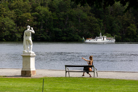 Stockholm, Sweden - July 13, 2019: A woman sitting on a bench next to a statue and water taking a selfie in the wonderful surroundings called Drottningholmのeditorial素材