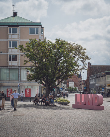 Lund, Sweden - August 4, 2020: People eating ice cream on a bench under a tree next to the city name...