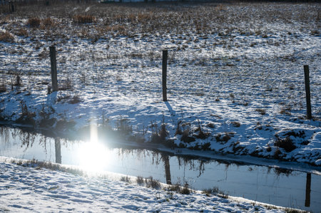 Sunlight reflection in rural water stream next to pasture fence all covered in snowの写真素材