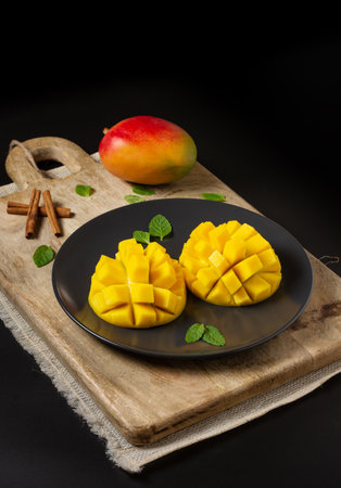 Mango cut in half and cubed on a wooden board, a whole mango and cinnamon branches, black background with space for copyingの写真素材