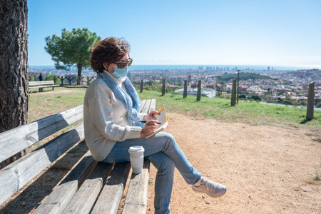 A woman a surgical mask, sitting on a bench writing in her notebook with the city in the backgroundの写真素材