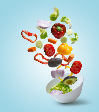 Sliced vegetables falling into bowl with salad on blue backgroundの写真素材