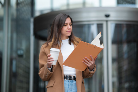 Businesswoman is holding a coffee and reading documents on a clipboard outside of an office building