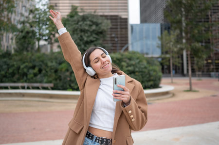 Carefree young woman enjoying music through headphones, holding smartphone and dancing in urban city...