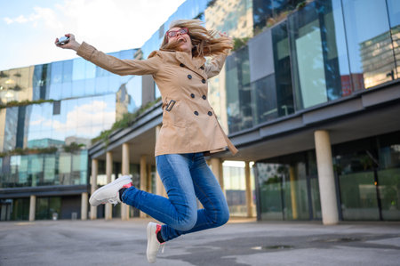 Excited businesswoman celebrating success by jumping in the air outside a modern office buildingの写真素材