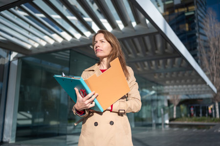 Businesswoman holding folders and looking away while standing outside modern office buildingの写真素材