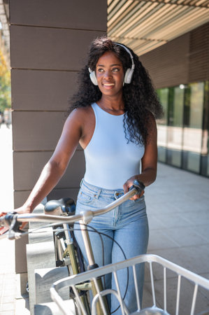 Stylish young woman is listening to music with headphones and holding her bicycle, enjoying a sunny day in the cityの写真素材