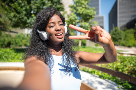 Happy young woman taking selfie while listening music with headphones and making victory sign with...