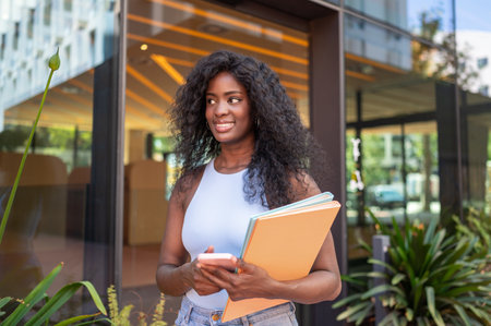 Cheerful black female student holding folders and using a smartphone while walking outside university buildingの写真素材