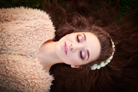 beautiful young woman With a fashionable make-up in a light cardigan and a wreath of flowers lying on grassの写真素材