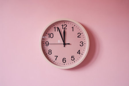 A round wall clock with black hands and numbers sits against a soft pink wall. The clock displays the time just before noon, creating a calm, inviting atmosphere.の素材