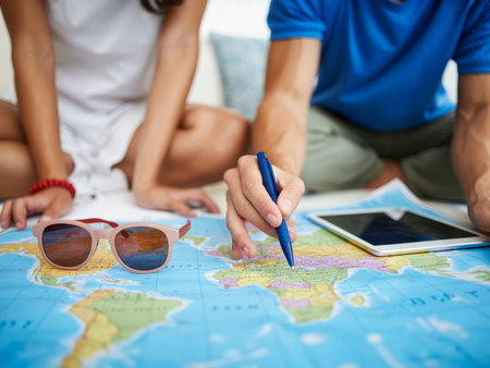 Two friends gather around a map laid out on the floor, planning their next travel adventure. One writes on the map with a pen while the other looks on, wearing stylish sunglasses.の素材