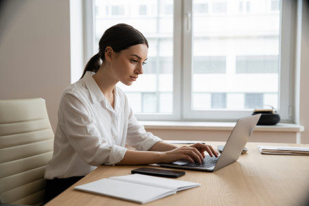 A focused woman in a white shirt types on her laptop at a modern office desk. Natural light from a large window brightens the space as she concentrates on her work.の素材