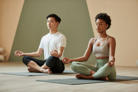 A young Asian man and a Black woman sit cross-legged in a calm indoor setting, practicing meditation together. They focus on their breath and maintain a tranquil atmosphere.の素材