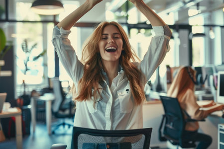 A young woman beams with joy, arms triumphantly raised in a gesture of victory, seated in an office setting.の素材