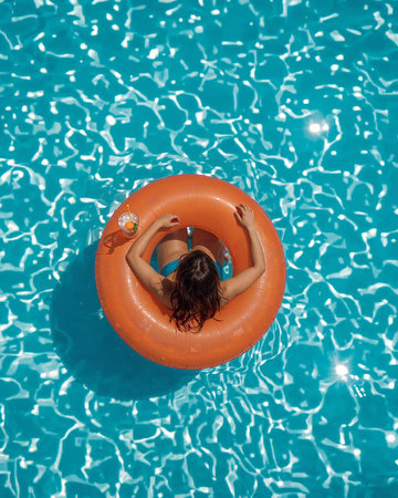 A woman floats peacefully in a bright orange pool float on a clear blue pool. She sips a refreshing drink while enjoying the warm sunshine.の素材
