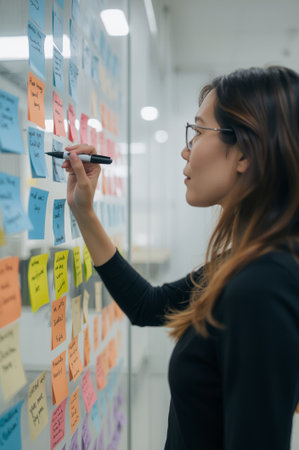 In a bright office, a professional Asian woman writes on colorful sticky notes attached to a glass board. She focuses on organizing ideas and planning projects during work hours.の素材