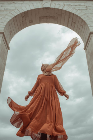 A woman wearing a long, flowing dress and a light scarf stands beneath a large stone arch. Her dress billows softly in the breeze as clouds drift by, creating a serene atmosphere.の素材