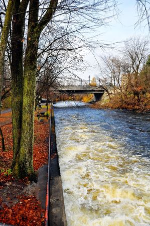 Alongside the water canal in the city during fall with leafless trees and bridge on a cloudy day.の写真素材