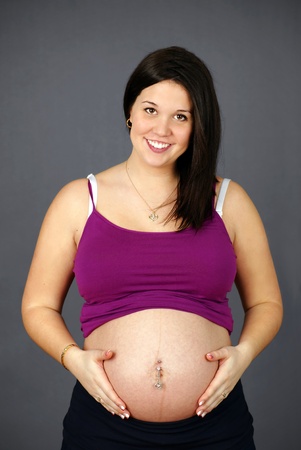 Studio shot portrait of a naturally beautiful, happy and healthy pregnant young woman holding her belly and smiling over grey background.の写真素材