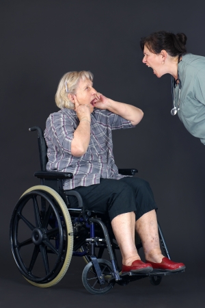 Elder abuse concept with a senior woman in a wheelchair crying and covering her ears as a middle age nurse or other health care worker is yelling at her.の写真素材
