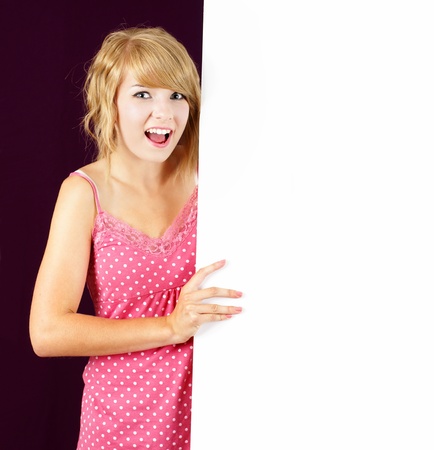 Cute and friendly smiling young blond teenager girl holding and pointing at a white blank sign, placard or billboard.の写真素材