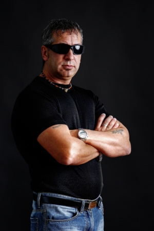 Somber tough guy with tatoos and black sunglasses, arms crossed, looking at camera, studio shot over black.の写真素材