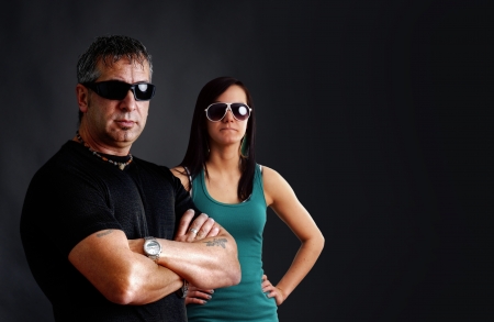 Biker couple: tough guy with tatoos and black sunglasses, arms crossed, looking at camera with pretty young brunette woman, studio shot over black.の写真素材