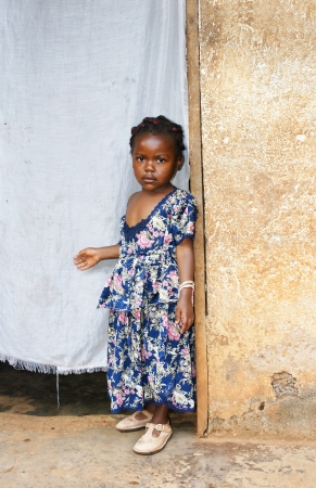 Cute but serious little black African girl in pink sunday dress next to her home door made of fabric; third world or developing country concept.の写真素材