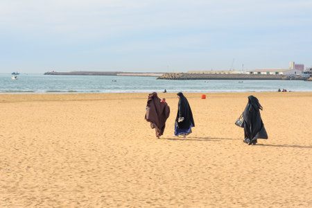 Agadir. Morocco - December 23, 2016: Muslim women in black long gowns walking on the beachのeditorial素材