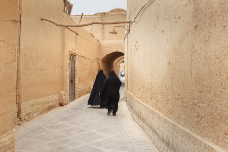 Yazd, Iran - March 23, 2018: Two Muslim women, dressed in black chador walk on the narrow street of old adobe Yazd cityのeditorial素材
