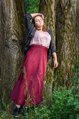 Brunette girl near tree in park. Girl in black jacket and red skirt with long hair on the street of...