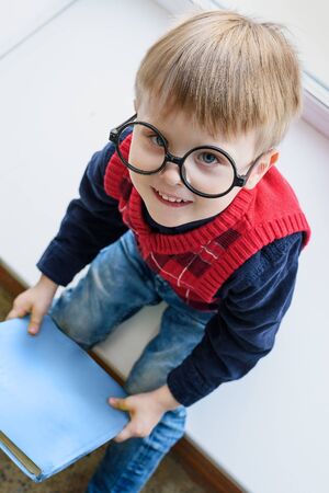 Portrait of little boy in glasses with book in library on windowの写真素材